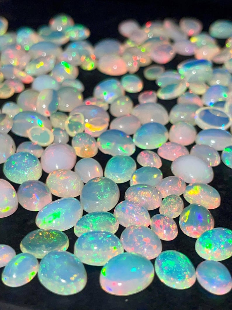 May include: A collection of oval-shaped opals in various sizes. The opals display a range of colors, including white, blue, green, and pink, with iridescent flashes. The gemstones are scattered across a dark surface.