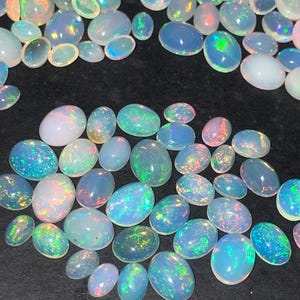 May include: A collection of oval-shaped opals in various sizes, displaying a range of colors including white, blue, green, and pink. The opals have a shimmering, iridescent quality, with flashes of color across their surfaces. The gemstones are scattered on a dark surface.