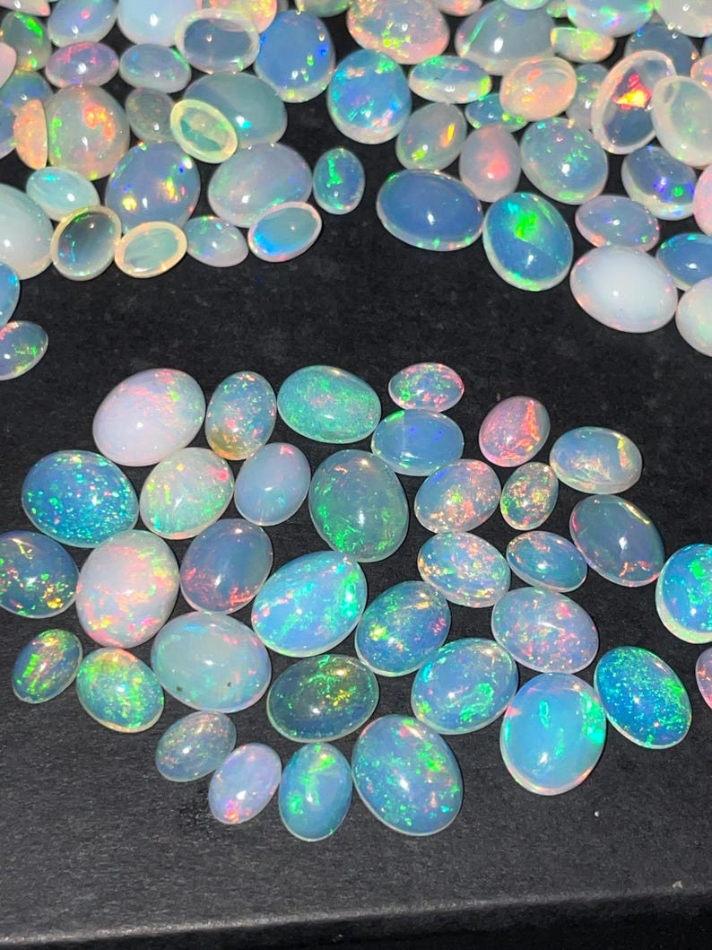 May include: A collection of oval-shaped opals in various sizes, displaying a range of colors including white, blue, green, and pink. The opals have a shimmering, iridescent quality, with flashes of color across their surfaces. The gemstones are scattered on a dark surface.