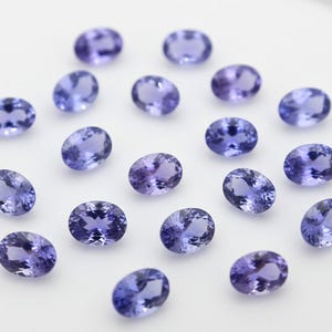 Può includere: Una collezione di gemme ovali, blu-viola, disposte su una superficie bianca. Le gemme presentano una gamma di sfumature, dal viola intenso al blu chiaro. Le pietre lucide riflettono la luce, evidenziando la loro chiarezza e brillantezza.