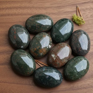May include: A collection of polished, oval-shaped gemstones in shades of deep green, brown, and red. The stones are arranged in a circular pattern on a wooden surface, with a small sprig of dried greenery in the background. The gemstones have a glossy finish.