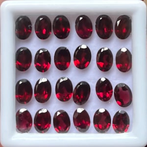 Puede incluir: Una colección de gemas ovaladas de color rojo intenso dispuestas en una bandeja blanca. Las gemas tienen una superficie pulida y reflectante, mostrando su rico color y claridad. La bandeja está dividida en filas.