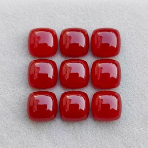 May include: Nine polished, vibrant red cabochons arranged in a three-by-three grid. Each gemstone features a smooth, rounded square shape with a high-gloss finish that reflects light, creating a bright white highlight on the surface of each piece.