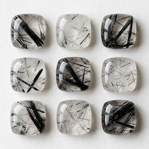 Puede incluir: Nueve piedras preciosas de cuarzo rutilado pulidas de forma cuadrada, dispuestas en una cuadrícula de tres por tres. Cada piedra transparente presenta inclusiones negras distintivas en forma de aguja, que varían en densidad y orientación.