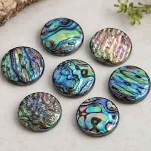 May include: Seven circular abalone shell buttons featuring iridescent surfaces with swirling patterns of blue, green, purple, and pink. Each button has a polished finish and displays unique natural color variations across its round, flat shape.