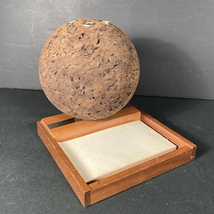 May include: A spherical cork object with metal rings on top, resting on a wooden tray with a light-colored insert. The cork has a textured, brown appearance. The wooden tray has a rectangular shape.