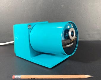 May include: A bright turquoise electric pencil sharpener with a cylindrical body and a metallic silver circular opening. The brand name Panasonic is printed in white on the front. A standard wooden pencil is placed in front for scale reference.