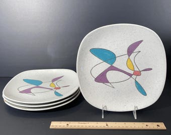 May include: A set of four square ceramic plates with rounded corners, featuring a speckled white finish and an abstract design of black lines with blue, purple, and yellow shapes. A wooden ruler shows the plates are approximately 10 inches wide.