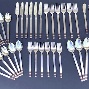 May include: A collection of vintage silverware, including spoons, forks, knives, and butter knives. The stainless steel utensils have a simple, elegant design with a brown band detail near the base of the handles. The arrangement suggests a formal dining setting.