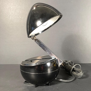 May include: A vintage black desk lamp with a rounded shade and base. The lamp features a chrome-colored adjustable arm and a power cord. The shade is open, revealing the light bulb. The lamp is designed to sit on a desk or table.