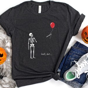 May include: A dark heather gray short-sleeve t-shirt featuring a white graphic of a skeleton looking at a red balloon floating away, with the text "Well, shit..." printed below the skeleton. The shirt has a crew neckline and a standard fit.