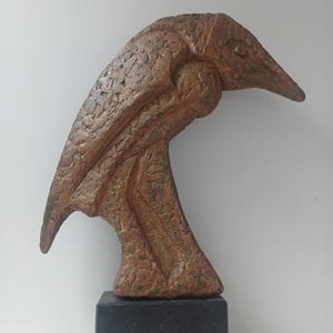 May include: A bronze-coloured sculpture of a bird's head and neck, mounted on a black rectangular base. The sculpture has a textured surface and a long, pointed beak. The bird's head is tilted downwards, giving it a graceful appearance.