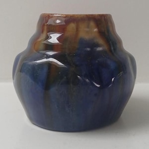 Peut inclure: Un petit vase en céramique fait à la main, de forme unique et à la finition brillante. Le vase présente un mélange de couleurs, notamment le bleu profond, le marron et des touches de vert. Le design est organique et texturé.