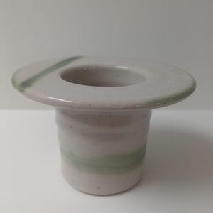 Może przedstawiać: Ceramiczny świecznik z szerokim, płaskim rondem i cylindryczną podstawą. Świecznik jest głównie w kolorze złamanej bieli z zielonym paskiem wokół podstawy i zielonym akcentem na rondzie. Projekt jest prosty i nowoczesny.