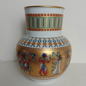 Peut inclure: Vase en céramique blanche et dorée sur le thème égyptien. Le vase présente des illustrations détaillées de personnages et de hiéroglyphes en or, rouge, bleu et turquoise. Le haut et la base ont des motifs décoratifs, et le bord est orné d'or.