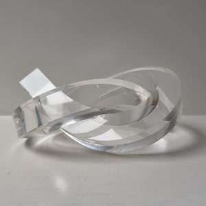 Puede incluir: Una escultura abstracta transparente con un diseño en forma de nudo. El material transparente refleja la luz, creando reflejos y sombras. La escultura está sobre una superficie blanca, con una etiqueta blanca.