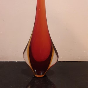 Peut inclure: Un vase en verre en forme de goutte d'eau, de couleur orange, rouge, jaune et blanc. Il présente un col étroit et une base large et arrondie. Le vase est posé sur une surface sombre.