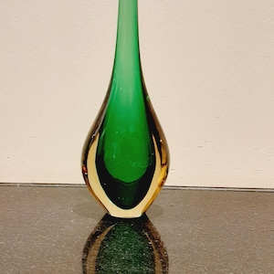Peut inclure: Un vase en verre en forme de goutte d'eau, avec un intérieur vert et un extérieur ambré transparent. Le vase a un long col fin et repose sur une surface réfléchissante. Le vase mesure environ 30 cm de haut.