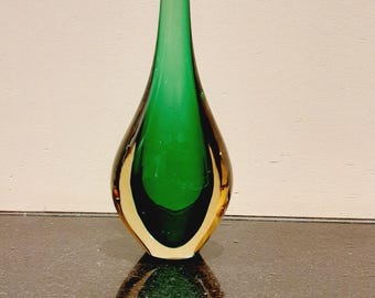 May include: A tall, teardrop-shaped glass vase with a green interior and a clear, amber-colored exterior. The vase has a long, slender neck and sits on a reflective surface. The vase is approximately 12 inches tall.