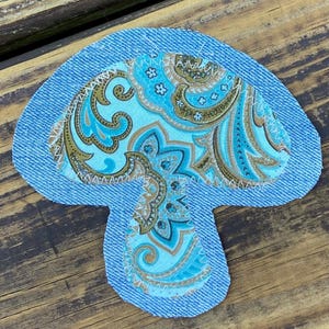 May include: A mushroom-shaped fabric patch featuring a blue denim border and a central section of light blue fabric with a brown and teal paisley pattern. The two fabric layers are joined together with visible white zigzag stitching along the edges.