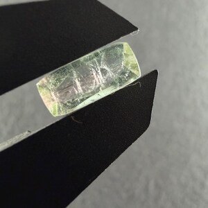May include: A rectangular, translucent light green gemstone held between the black metal jaws of a caliper. The stone features internal inclusions and a subtle pinkish hue visible within its crystalline structure, showcasing a polished surface.