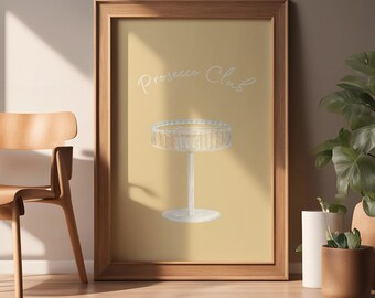 May include: A framed print featuring a champagne glass illustration with the text "Prosecco Club" in a handwritten style. The artwork has a light yellow background and is displayed in a wooden frame. A wooden chair and potted plants are in the background.