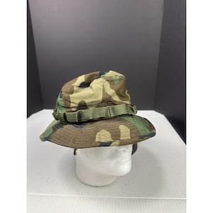 May include: Camouflage pattern boonie hat with a wide brim and a green band around the crown. The hat is made of a durable fabric and is designed for outdoor use. The hat is placed on a white mannequin head.