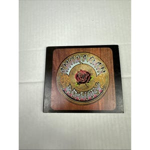 May include: A wooden coaster with a circular design featuring a red rose at the center. The design includes ornate gold and teal accents, with the words "Summer Camp" in a stylized font. The coaster is set against a dark border.