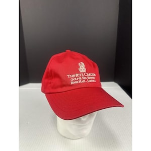 May include: A red baseball cap with a curved brim. The cap features white text that reads "THE RITZ-CARLTON GOLF & SPA RESORT ROSE HALL, JAMAICA" and a logo. The cap is displayed on a white mannequin head.