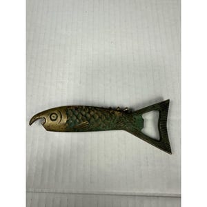 May include: A metallic bottle opener shaped like a fish with a weathered bronze and green patina finish. The design features engraved scale details along the body, a circular eye, and a functional tail section that serves as the opening mechanism.