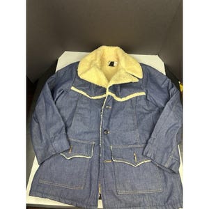 May include: A blue denim jacket featuring a cream-colored sherpa fleece collar and lining. The jacket includes a front button closure, two large lower pockets with pointed flaps, and a horizontal yoke seam across the chest with sherpa trim.