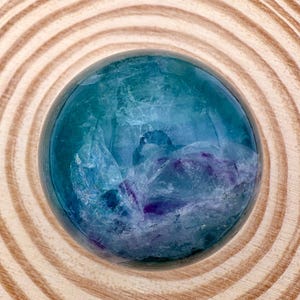 May include: A polished fluorite sphere, displaying blue and purple hues. The crystal ball rests on a wooden surface with concentric circular patterns. The sphere's interior has a cloudy, textured appearance.
