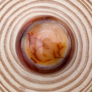May include: A polished, round gemstone with a translucent, amber-coloured centre and reddish-brown veining. The stone is set within a circular, wooden frame, showcasing the natural grain of the wood. The overall design is simple and elegant.