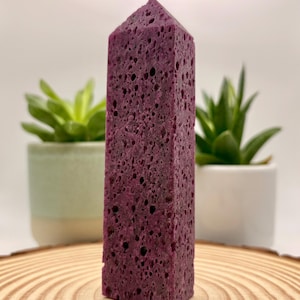 May include: A tall, six-sided crystal tower with a deep purple hue and a porous, textured surface. The stone features a pointed, faceted top and a rough, pitted exterior throughout its length, standing upright on a flat, circular wooden base.