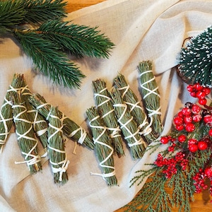 Könnte beinhalten: Anordnung von grünen Salbeibündeln, gebunden mit weißem Faden, zusammen mit Zweigen von immergrünen Pflanzen und roten Beeren. Die Bündel sind auf einem neutralen Stoff angeordnet und schaffen eine natürliche und festliche Ästhetik.