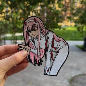 May include: An embroidered patch featuring an anime character with long pink hair, two small red horns, and a white bodysuit with red accents. The character is winking and making a peace sign gesture with one hand, outlined in a thick black border.