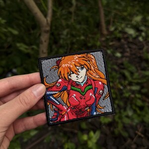 May include: A square embroidered patch featuring an anime character with long orange hair and blue eyes, wearing a red and blue plugsuit with the number 02 visible on the chest. The background of the patch is a textured grey pattern with a black border.
