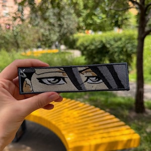 May include: A rectangular embroidered patch featuring a stylized graphic illustration of two eyes with dark hair strands falling across the forehead. The design uses shades of grey, black, and tan thread with a dark grey border on a fabric base.