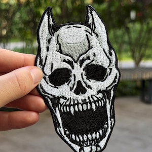May include: An embroidered patch featuring a stylised white skull with black accents and pointed ear-like protrusions at the top. The skull has deep black eye sockets and a wide, open mouth revealing detailed teeth, all outlined in a thick black border.
