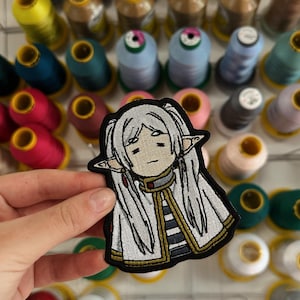 Puede incluir: Un parche bordado que presenta un personaje de anime estilizado con cabello largo blanco, orejas puntiagudas y una capa blanca con ribete dorado. El personaje tiene una expresión neutral y está representado en estilo chibi con un borde negro.