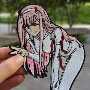 May include: An embroidered patch featuring an anime character with long pink hair, red horns, and green eyes. The character wears a white suit with red accents and is making a peace sign gesture with one hand. The patch has a black border.