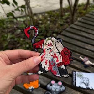 May include: An embroidered patch featuring a character wearing a white hooded cloak with red blood-like splatters, holding a curved scythe in one hand and a knife in the other, with black, red, and white thread details throughout the design.