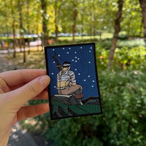Puede incluir: Un parche bordado rectangular que muestra una figura sentada sobre una roca frente a un cielo nocturno azul oscuro estrellado. La figura lleva vendas claras en el torso y la cabeza, con una gran espada a la espalda, sobre un paisaje verde.