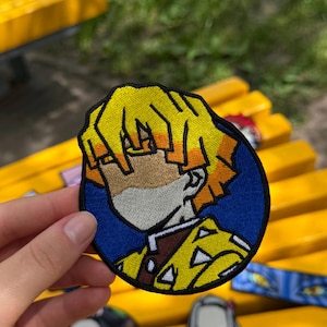 Könnte beinhalten: Ein kreisförmiger gestickter Aufnäher mit einem stilisierten Anime-Charakter mit leuchtend gelbem Haar, einem gebräunten Gesicht und einem dunkelbraunen Kragen vor einem einfarbigen blauen Hintergrund mit einem dicken schwarzen Rand.