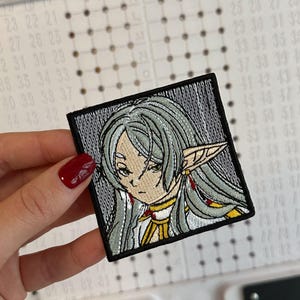 May include: A square embroidered patch featuring an anime-style character with long, light grey hair and pointed ears. The character has a serious expression, wearing a white and gold garment with red accents, all enclosed within a thick black border.