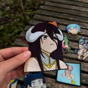 May include: An embroidered patch featuring an anime character with long dark hair, large yellow eyes, and prominent curved horns. The character wears a light-colored garment with gold accents. The patch has a defined black border and detailed stitching.