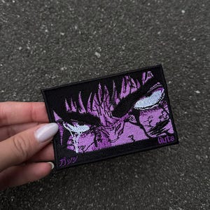 Puede incluir: Un parche rectangular bordado que presenta un primer plano estilizado de ojos intensos en tonos morados y negros. El diseño incluye reflejos blancos, el texto Guts en morado y los caracteres japoneses ガッツ en la esquina inferior izquierda.