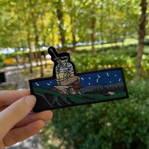 Puede incluir: Un parche rectangular bordado que muestra una figura con una gran espada atada a la espalda, sentada sobre una roca. La escena representa un cielo nocturno azul oscuro estrellado sobre un paisaje verde con raíces de árboles oscuras.