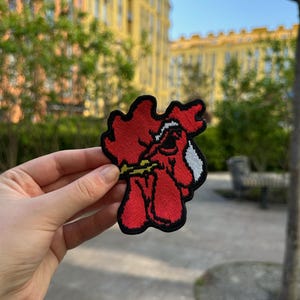 May include: An embroidered patch featuring a stylized red rooster head with a black outline, a white accent on the neck, and a yellow beak. The design is shaped with irregular edges and is held up against an outdoor background.