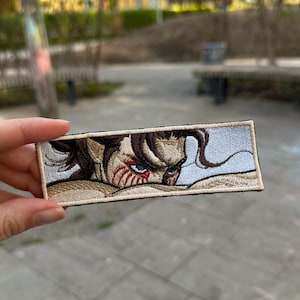 May include: A rectangular embroidered patch featuring a detailed anime-style character face with dark brown hair, green eyes, and red facial markings. The design is framed by a light beige border and a thin black outline on a white background.